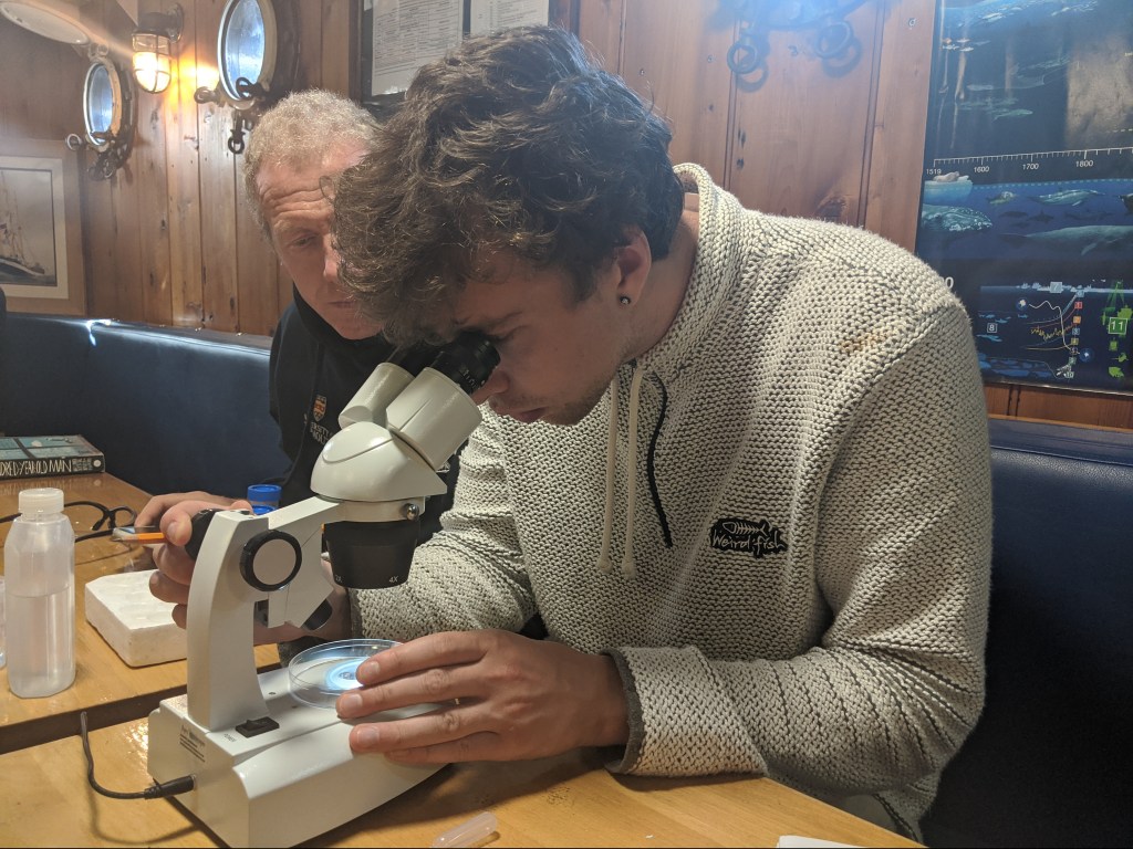 Young scientist searching for microplastics in a sample of seawater aboard the Pelican of London during the circumnavigation of the British Isles during the Darwin200.com voyage in summer 2020